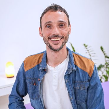 Smiling man in a denim jacket with brown shoulder patches, sitting indoors.
