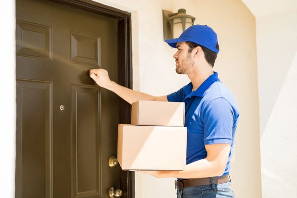 Delivery man, in a blue uniform, knocking on a door while holding boxes.