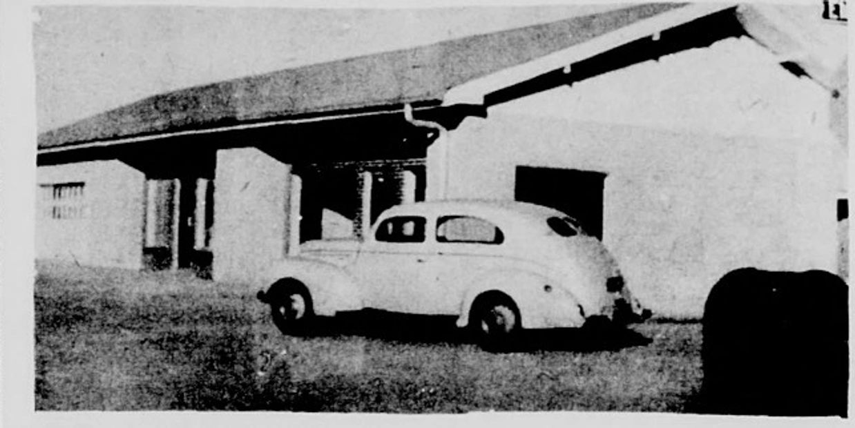 Vintage car parked outside a simple house with a sloped roof.