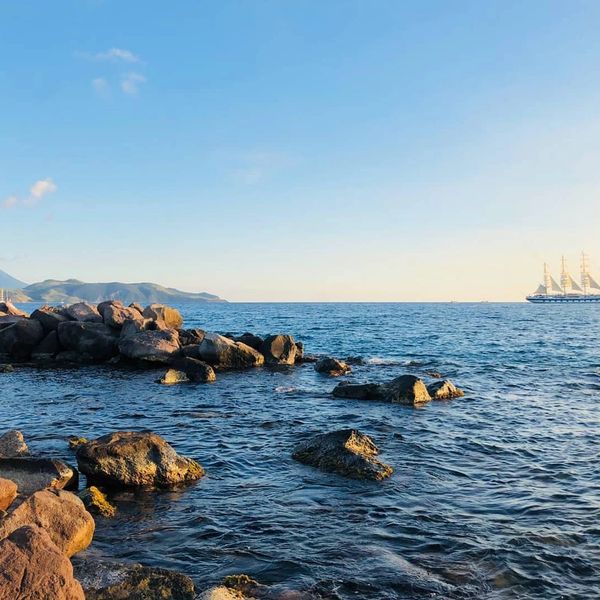 Rocky shoreline with a calm sea and a sailing ship in the distance under a clear sky.