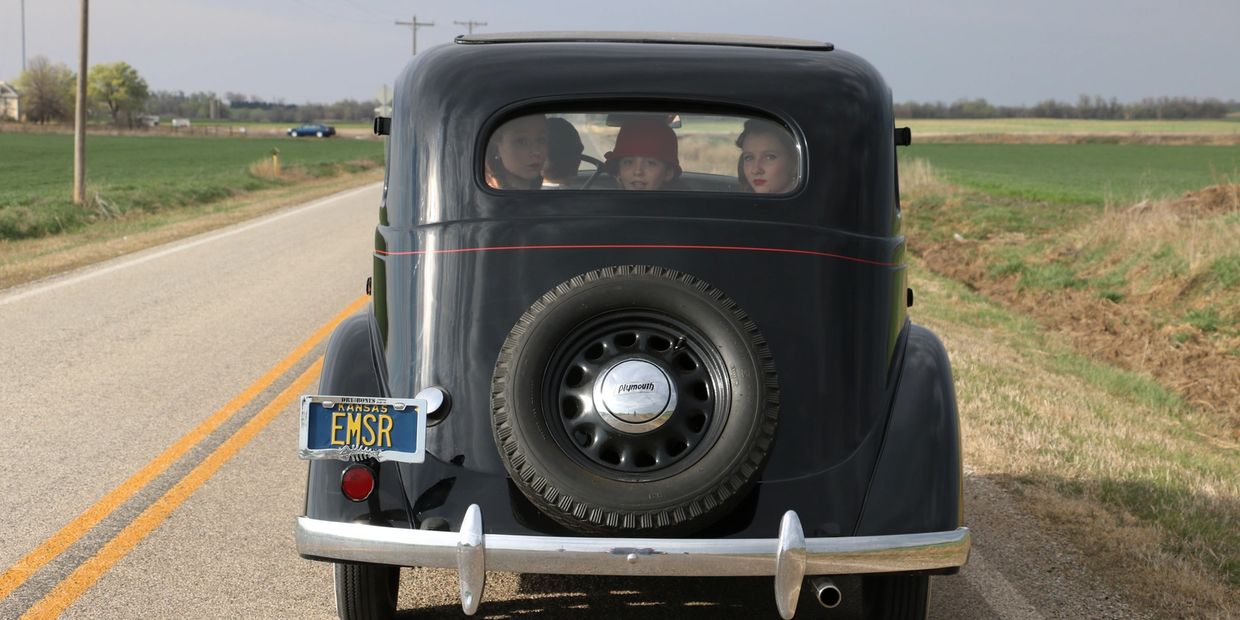 Three people inside an old black car on a country road.