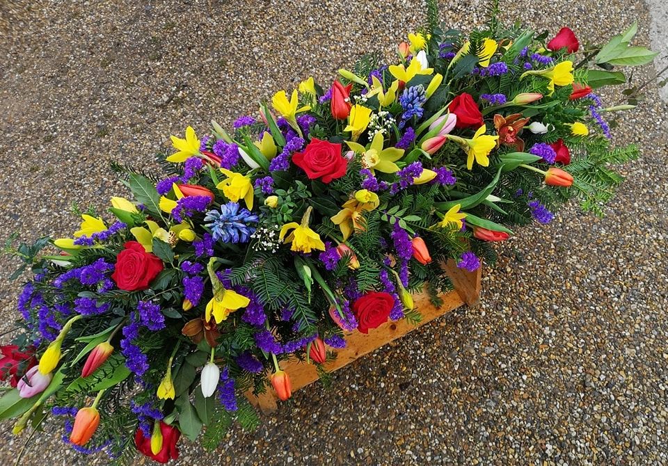A colorful coffin spray featuring a mix of bright yellow daffodils, red roses, and purple hyacinths.