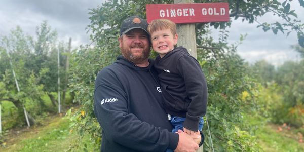 A man holding a smiling child in an apple orchard under a Ginger Gold sign.