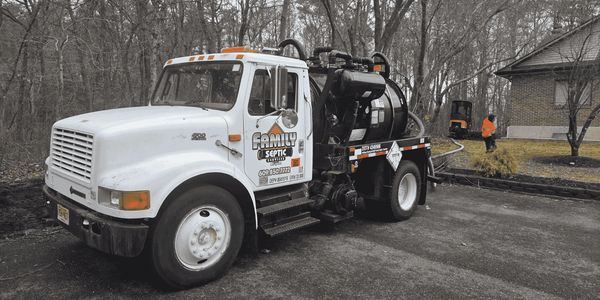 A white septic service truck parked near a house with a worker in the background.