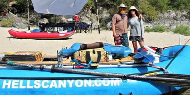Two people stand near a blue raft with gear on a sandy beach.
