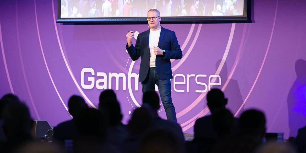 A man in a blazer giving a presentation on stage with a purple backdrop.