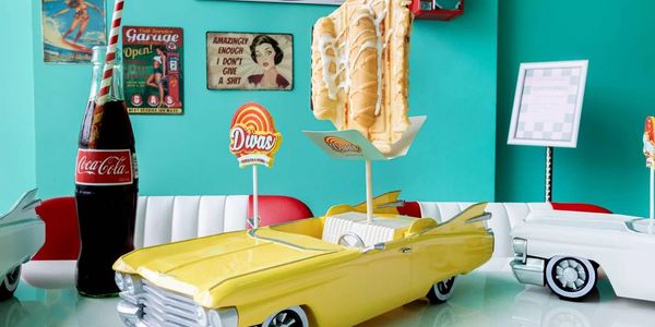 Retro diner scene with Cadillac Trays cars, a Coca-Cola bottle, and crepe on a stick.