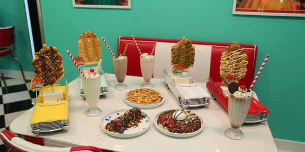 Retro diner table with milkshakes, ice cream, crepes, brunch, breakfast, lunch, desserts, and waffle pizza, pops in car-shaped holders.