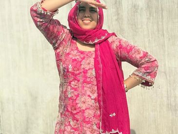A smiling woman in a pink floral dress and matching scarf shielding her eyes from the sun.