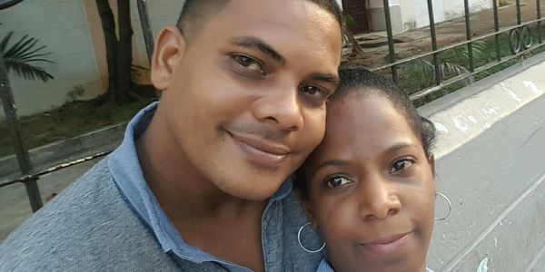 A smiling couple takes a close-up selfie outdoors near a concrete wall.