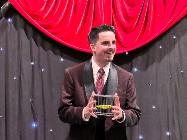 Man in a suit holding a small cage with a yellow bird on stage.