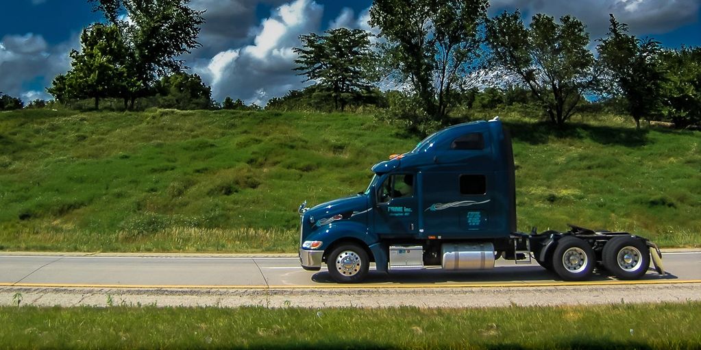 Blue semi-truck driving on a highway with green trees and grass in the background.