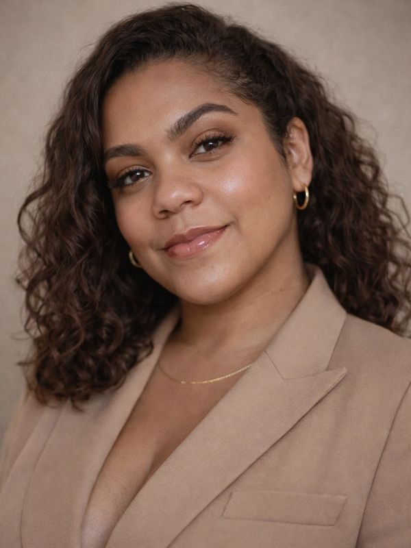 Confident woman with curly hair wearing a beige blazer and gold jewelry.