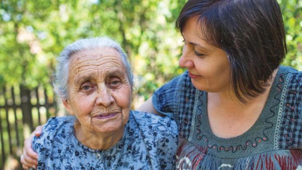An elderly woman and a younger woman share a tender moment outdoors.