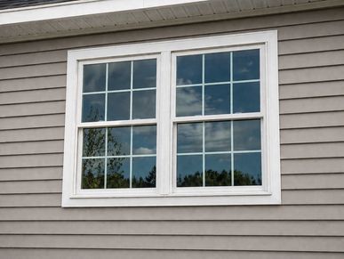 White-framed double window on a gray siding house reflecting the sky.
