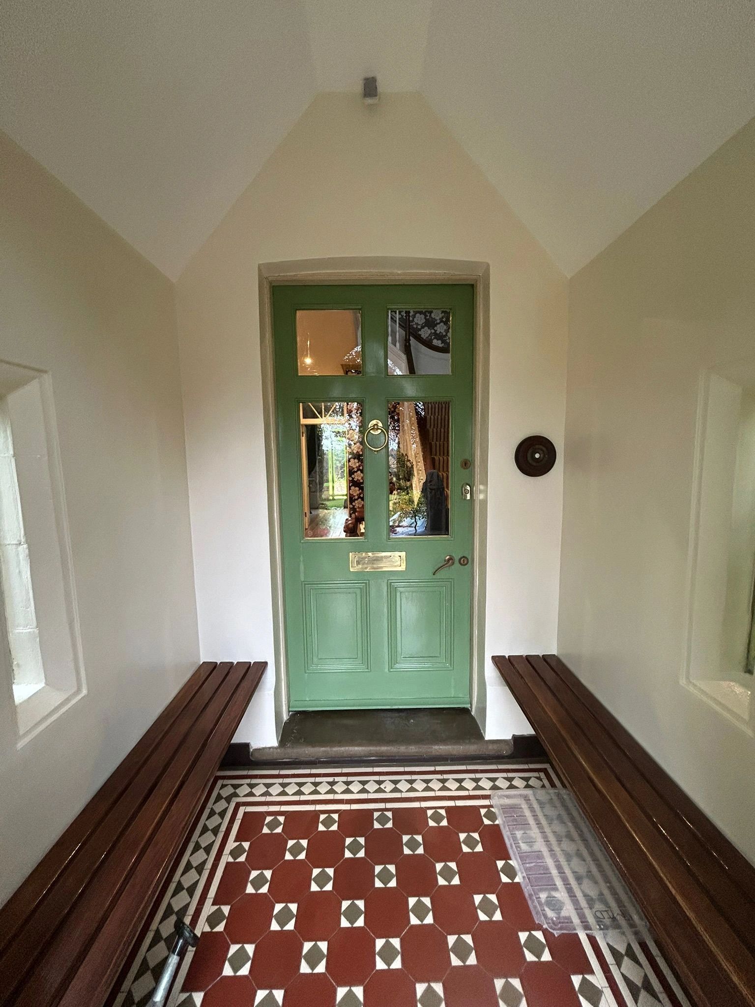 Green door with glass panels between wooden benches in a tiled hallway.