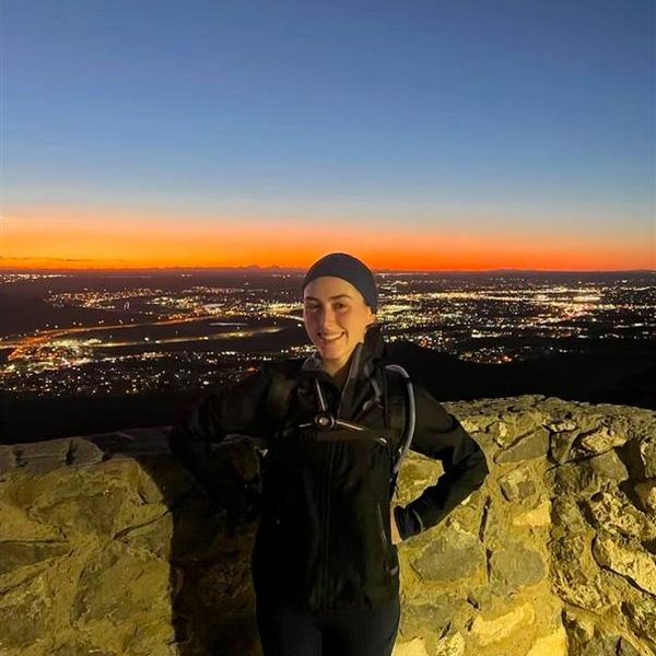 A woman stands smiling at sunset with city lights behind her.
