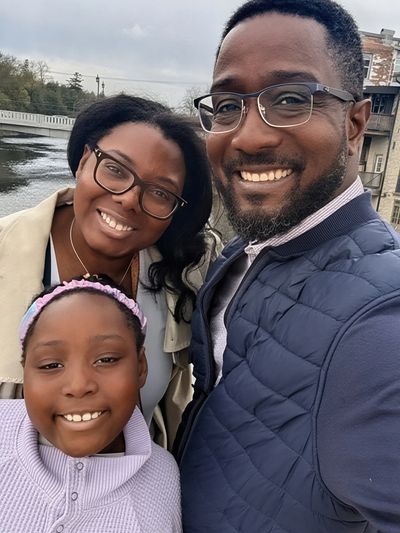 A happy family of three smiling outdoors by the water on a cloudy day.