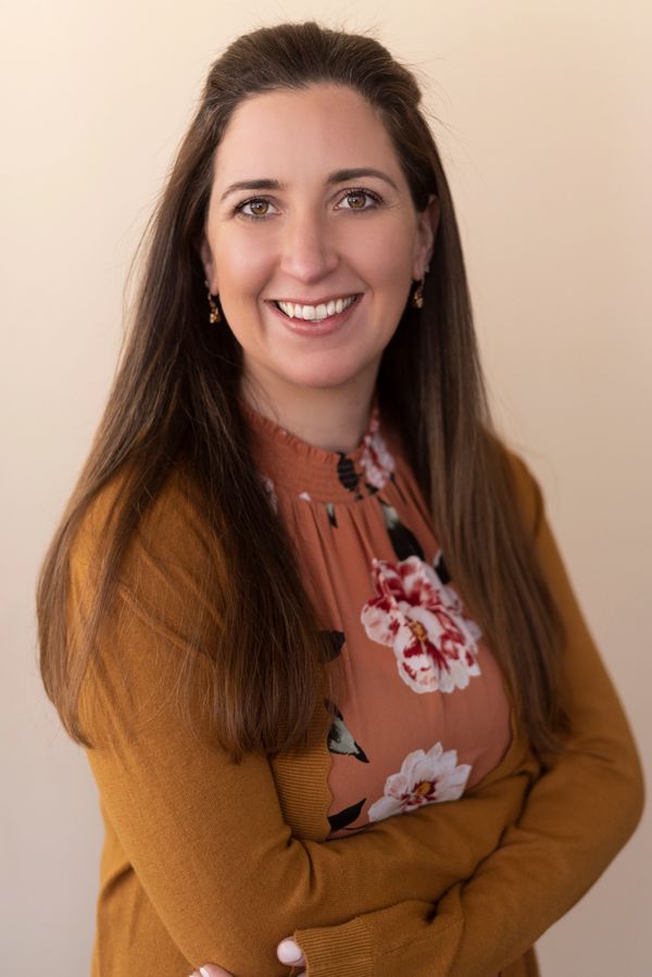 Smiling woman with long hair wearing a floral blouse and mustard cardigan.