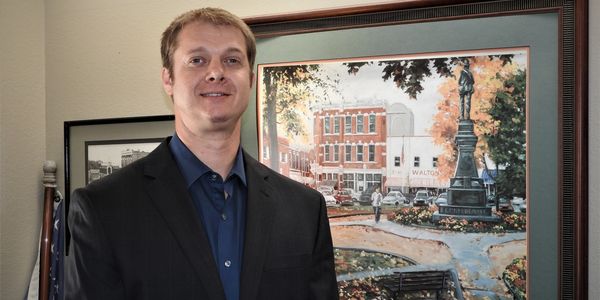 Man in a suit standing in front of a framed painting and an American flag.