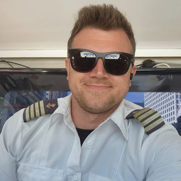 Pilot in uniform wearing sunglasses inside an aircraft cockpit.