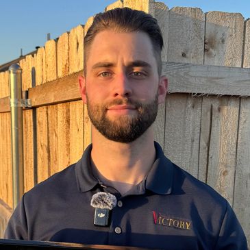 Man in navy blue Victory polo shirt stands outdoors near a wooden fence.