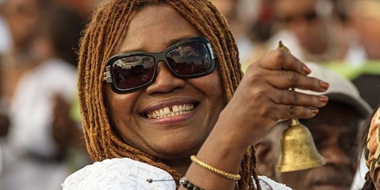 Smiling woman with dreadlocks ringing a brass bell at an outdoor event.
