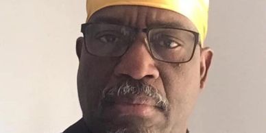 Man wearing glasses and a yellow headwrap looks serious.