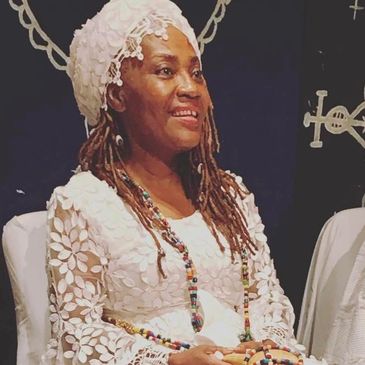A woman dressed in white traditional attire with a headwrap and beaded jewelry.