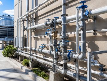 Industrial metal pipes and valves on a building exterior under sunlight.