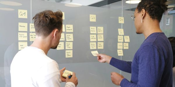 Two colleagues organize tasks on a glass board with sticky notes.