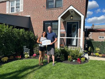 A happy couple stands in front of their new home holding a 'Sold' sign.