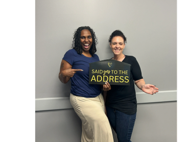Two women smiling, one holding a sign saying 'Said Yes to the Address.'