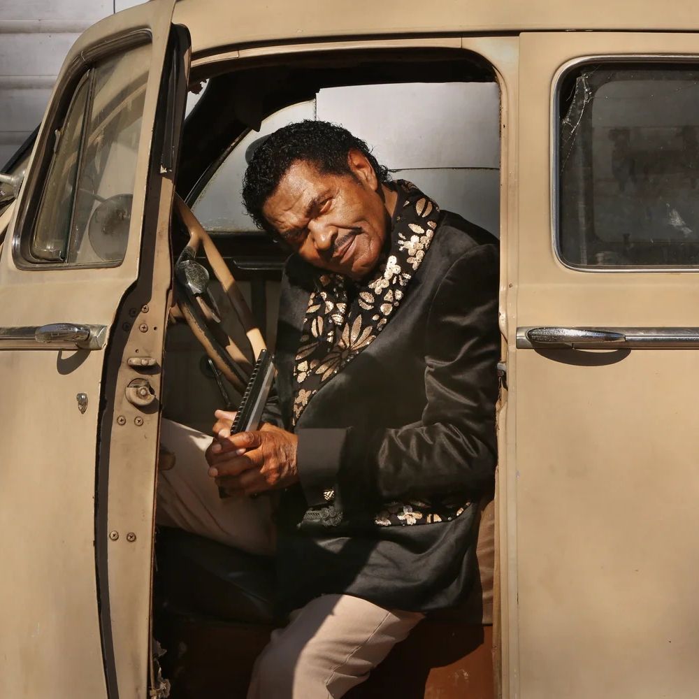 Man in vintage car holding a harmonica, dressed in a patterned jacket.