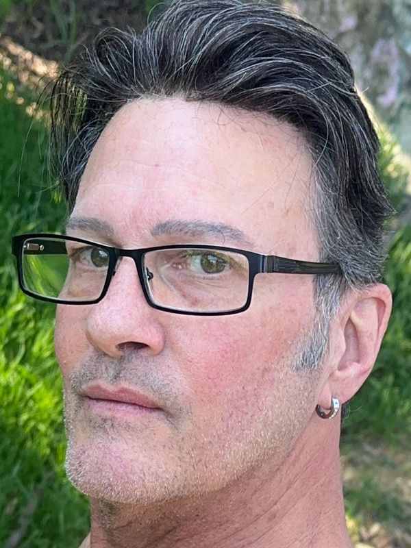 Close-up of a man with glasses and an earring outdoors.