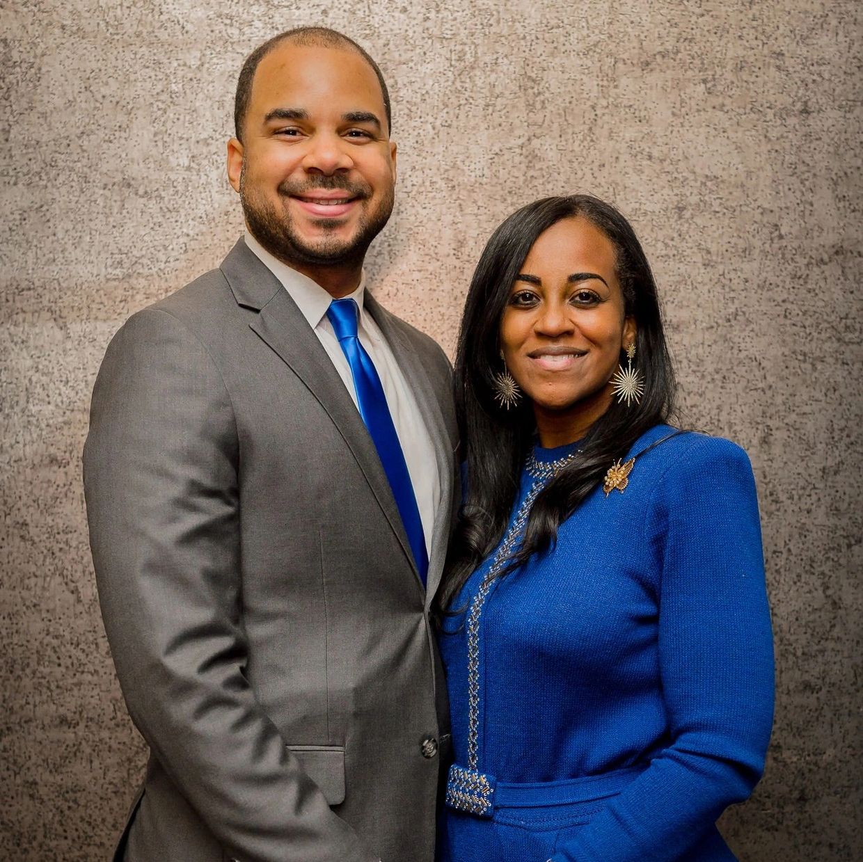 Smiling couple dressed formally against a textured wall.