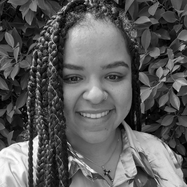 Smiling woman with braided hair and a cross necklace in front of leafy background.