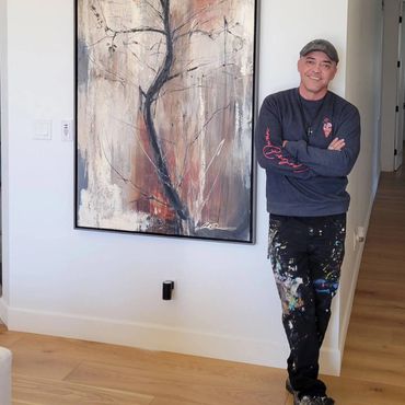 Man with paint-splattered pants stands next to abstract tree painting on wall.