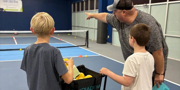Coach instructing two boys during tennis practice with a basket of balls.