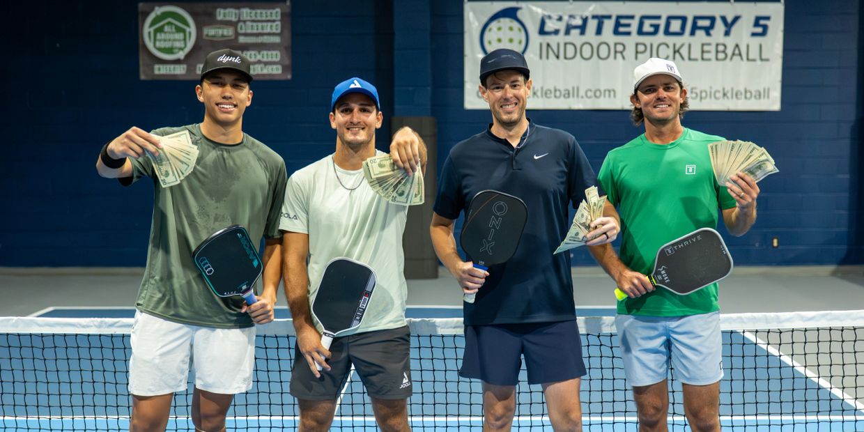 Four men holding pickleball paddles and cash prizes, smiling on an indoor court.
