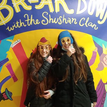 Two women with long hair wearing masks and black coats in front of a colorful event backdrop.