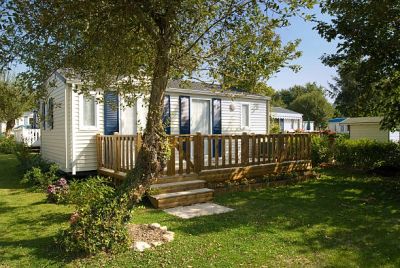 Small white mobile home with a wooden deck surrounded by green lawn and trees.