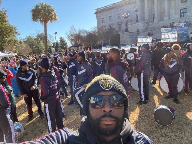 Man taking selfie with a marching band and crowd outside a government building.