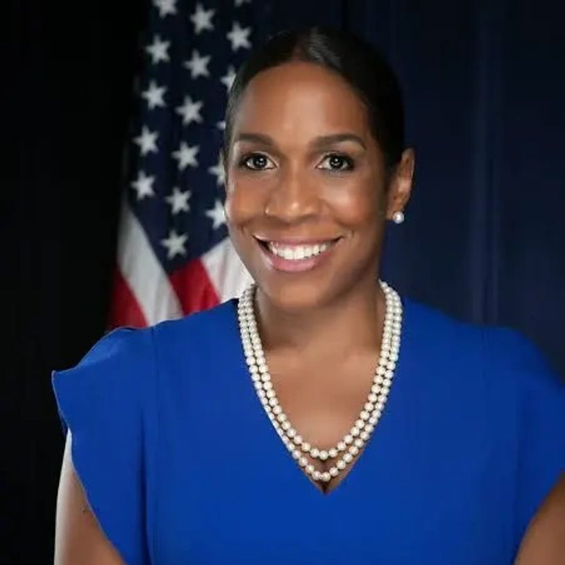 Professional woman in blue dress with pearl necklace smiling in front of American flag.