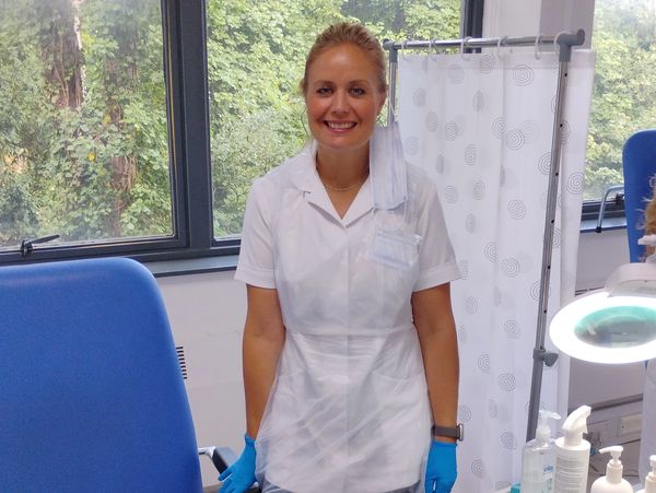 Smiling female healthcare worker in a clinic room with medical equipment and blue chair.