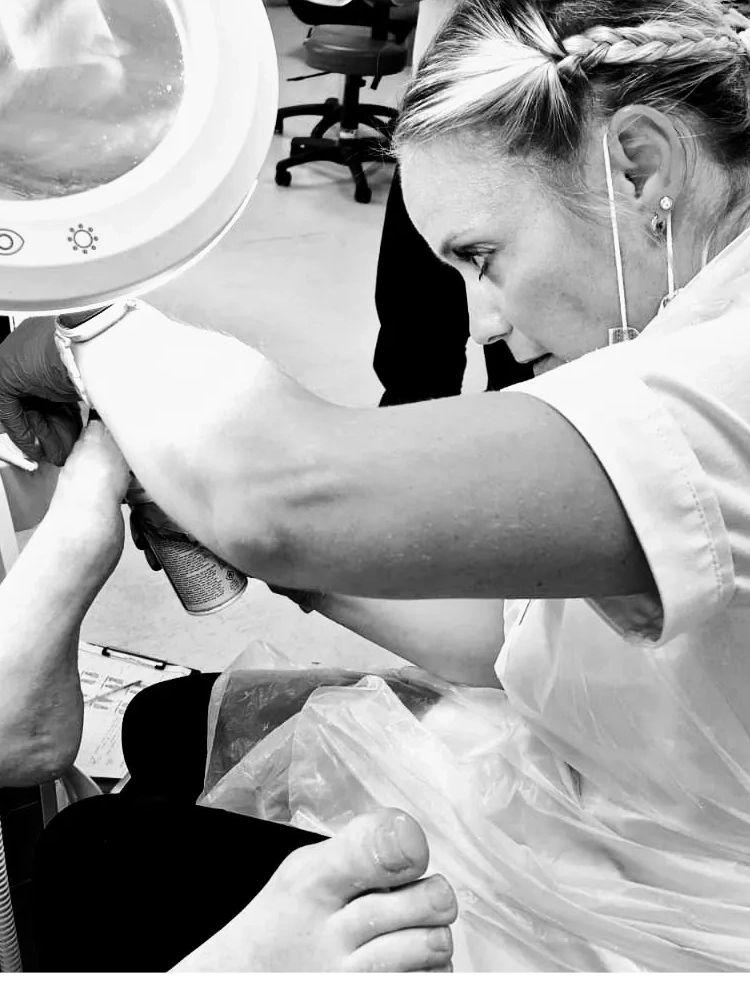 A foot health practitioner closely examines a patient's foot under a magnifying lamp in a clinical setting.