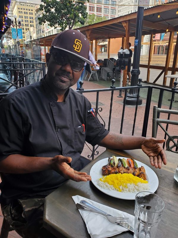Man in black chef coat presenting a plate of grilled chicken and rice outdoors.