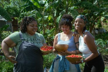 Tres mujeres sonrientes sostienen frutas recién cosechadas en un jardín.
