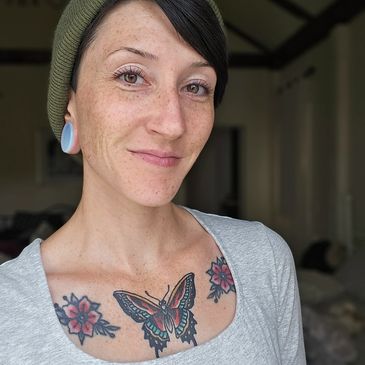 A woman with a butterfly tattoo and floral designs on her chest, smiling gently.