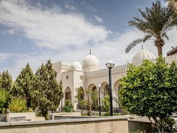 A white mosque with domes and arches surrounded by greenery under a partly cloudy sky.